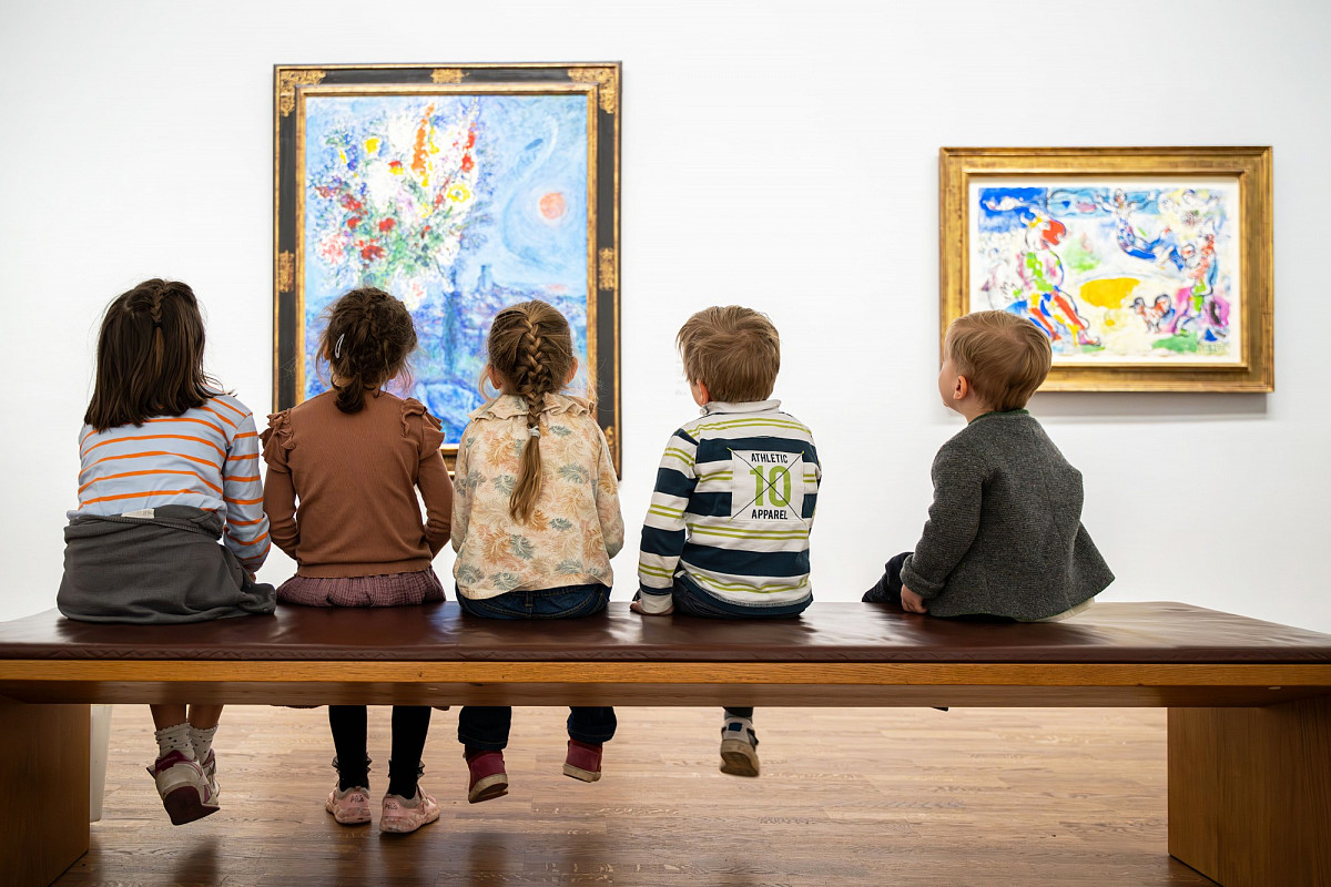Five children sit in a row on a bench in an exhibition. You can see their backs and the backs of their heads, in front of them the paintings Sleeping Woman with Flowers and The Great Circus by Marc Chagall | Photo © ALBERTINA Photo Studio