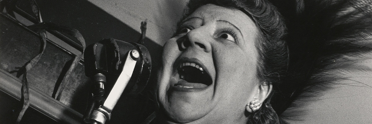 A woman in an evening dress holding a standing microphone up to her face. Her mouth is wide open. Credit: Lisette Model | Singer at the Cafe Metropole, New York City, 1946 | ALBERTINA Wien © 2025 Estate of Lisette Model, courtesy Lebon, Paris / Keitelman, Brussels