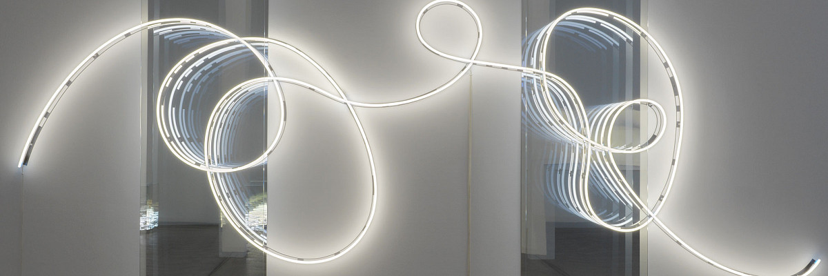 View into a dark exhibition room, illuminated only by a curved light installation in front of a mirror. | Brigitte Kowanz | Email 02.08.1984 03.08.1984, 2021 | ESTATE BRIGITTE KOWANZ © Estate Brigitte Kowanz / Bildrecht, Vienna 2025 | Photo: Peter Hoiss