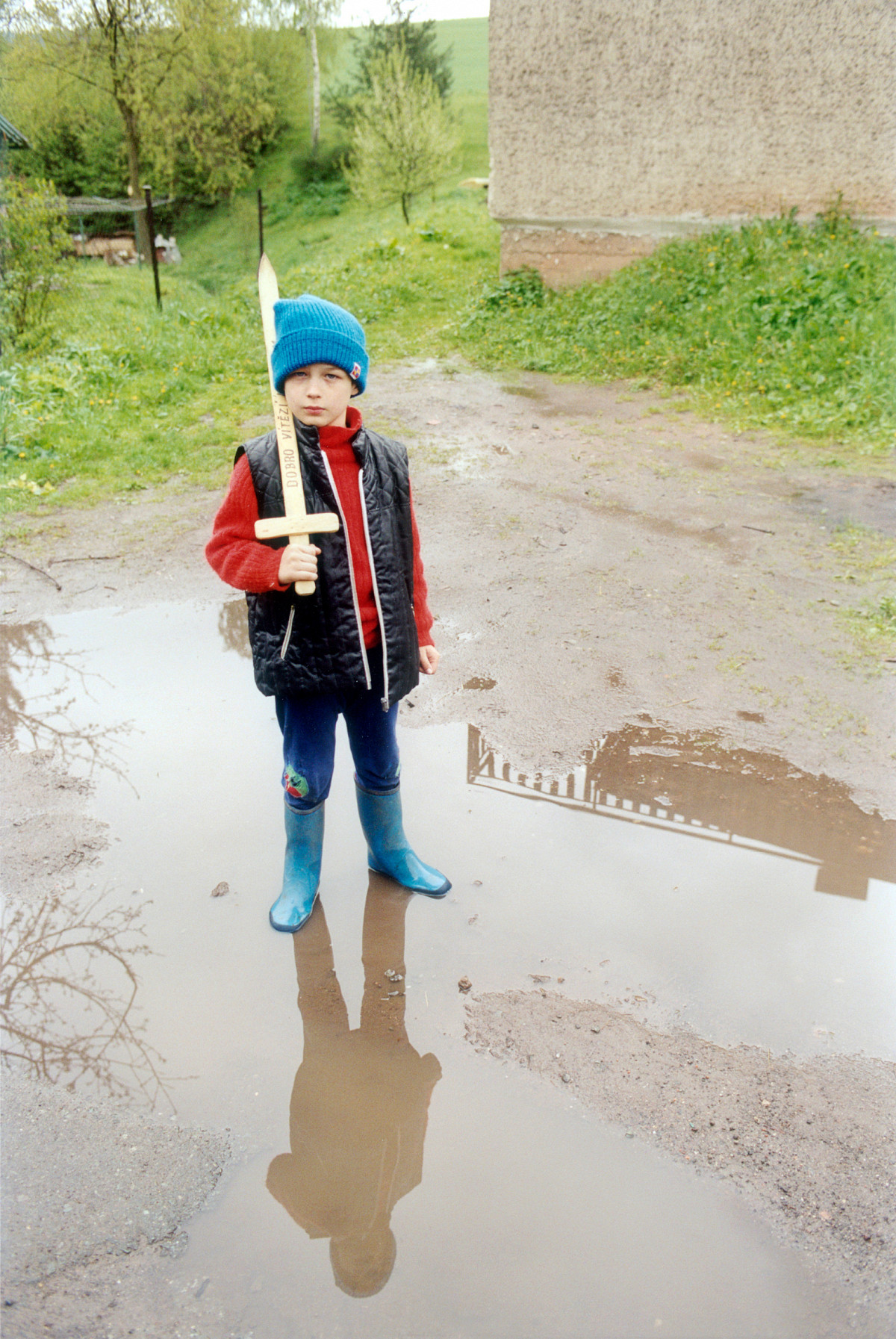 A little boy with a blue cap and blue wellies is standing in a puddle of rain, holding a wooden sword in his hand. He has leaned the sword over his shoulder.