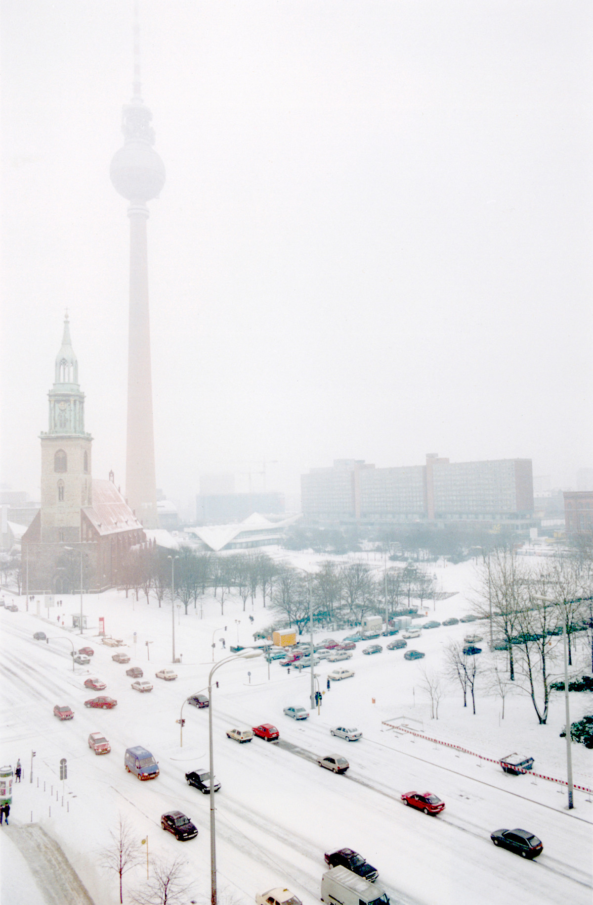 A snow-covered street with heavy traffic. In the background a church, a television tower and block buildings in the snow.