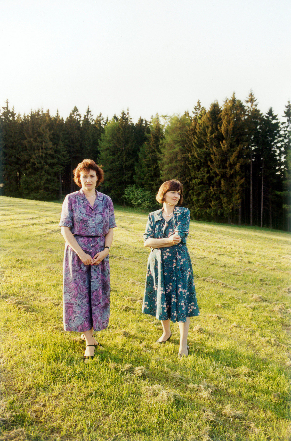 Two women stand next to each other in the field. The forest begins in the background. They are both wearing fitted, floral midi dresses with short sleeves. One woman looks into the camera, the other looks to the side. Both have chin-length brown hair.