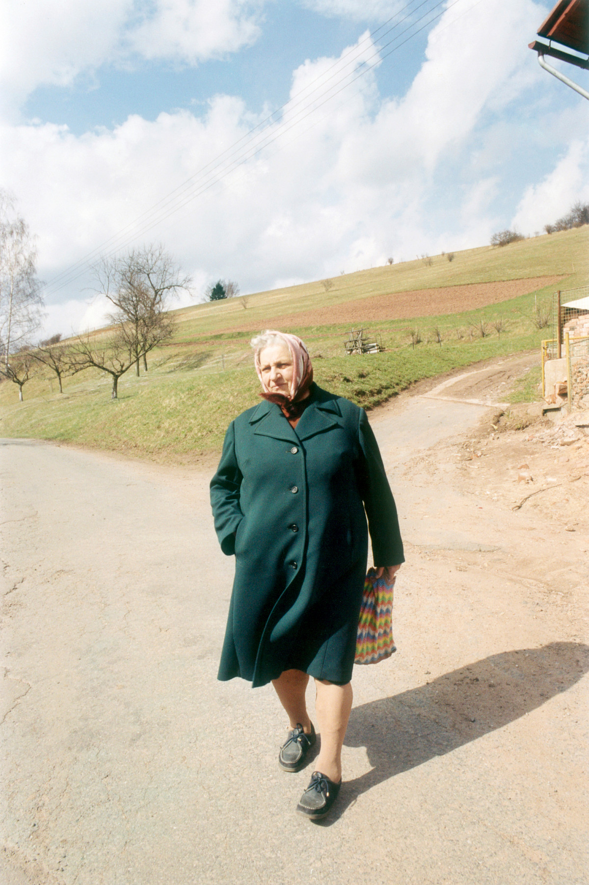 An old woman in the countryside. She is walking along a road, with fields and a few barren, small trees behind her. The woman is wearing a dark green coat and a headscarf. She is holding a colorful bag in her hand.