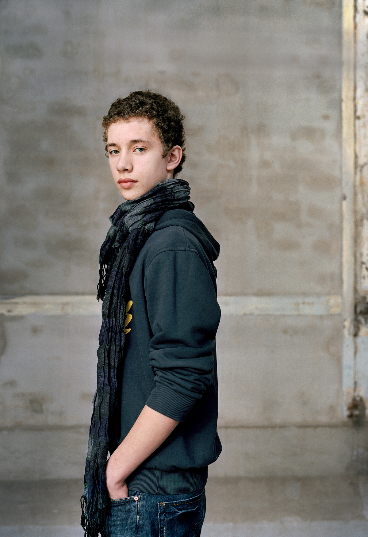 A teenage boy, standing sideways and looking into the camera. His hands are in his trouser pockets. He is wearing jeans, a hoodie and a scarf. A gray wall in the background.