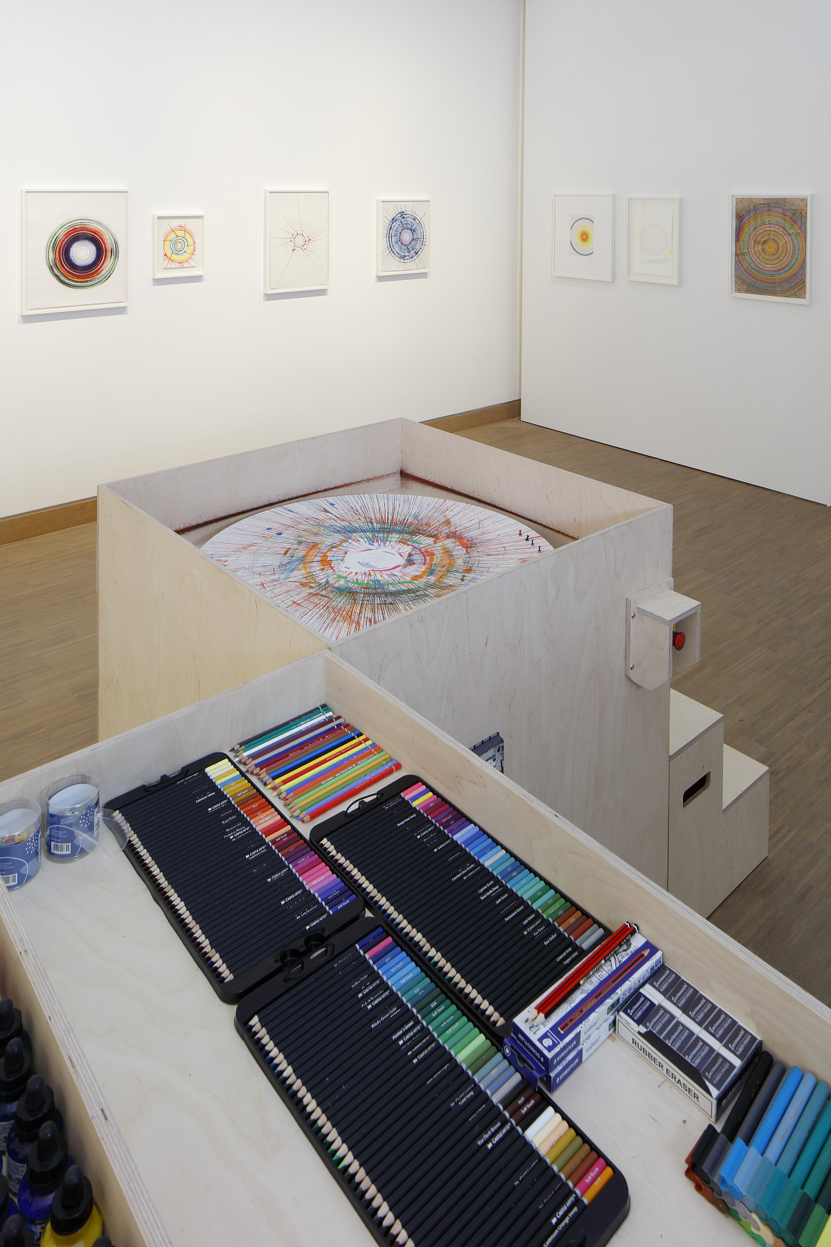 Photo of Damien Hirst's drawing machine with a large selection of colorful pens in a bright exhibition room with Hirst's ‘'Spin Drawings’' on the wall.