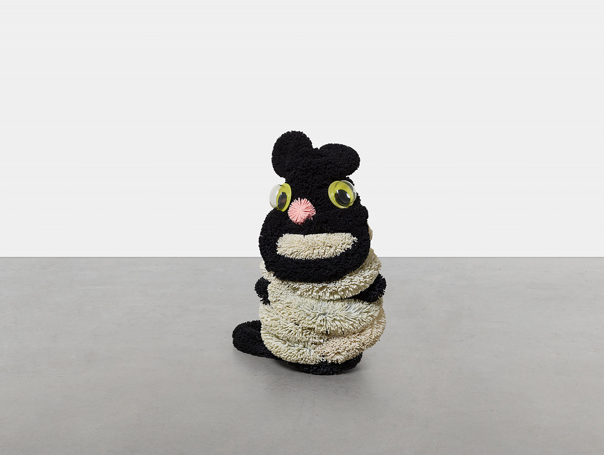 An upright sitting, black and beige plush figure reminiscent of a cat. With big googly eyes and a pink nose.