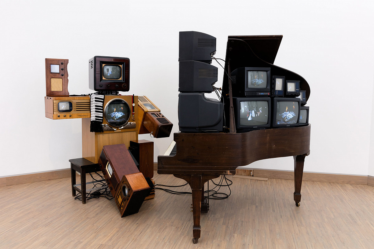 Photo of an installation: A piano with a stool. Several tube televisions are set up on the open grand piano. A robot-like figure, also constructed from old televisions and radios, sits on the stool.