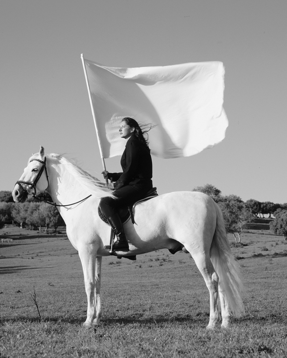 Black and white photograph of the artist sitting on a white horse and holding a large white flag. | Marina Abramović | The Hero, 2001, Courtesy of the Marina Abramović Archives, and Galeria Luciana Brito © Marina Abramović. Image courtesy of Marina Abramović Archives