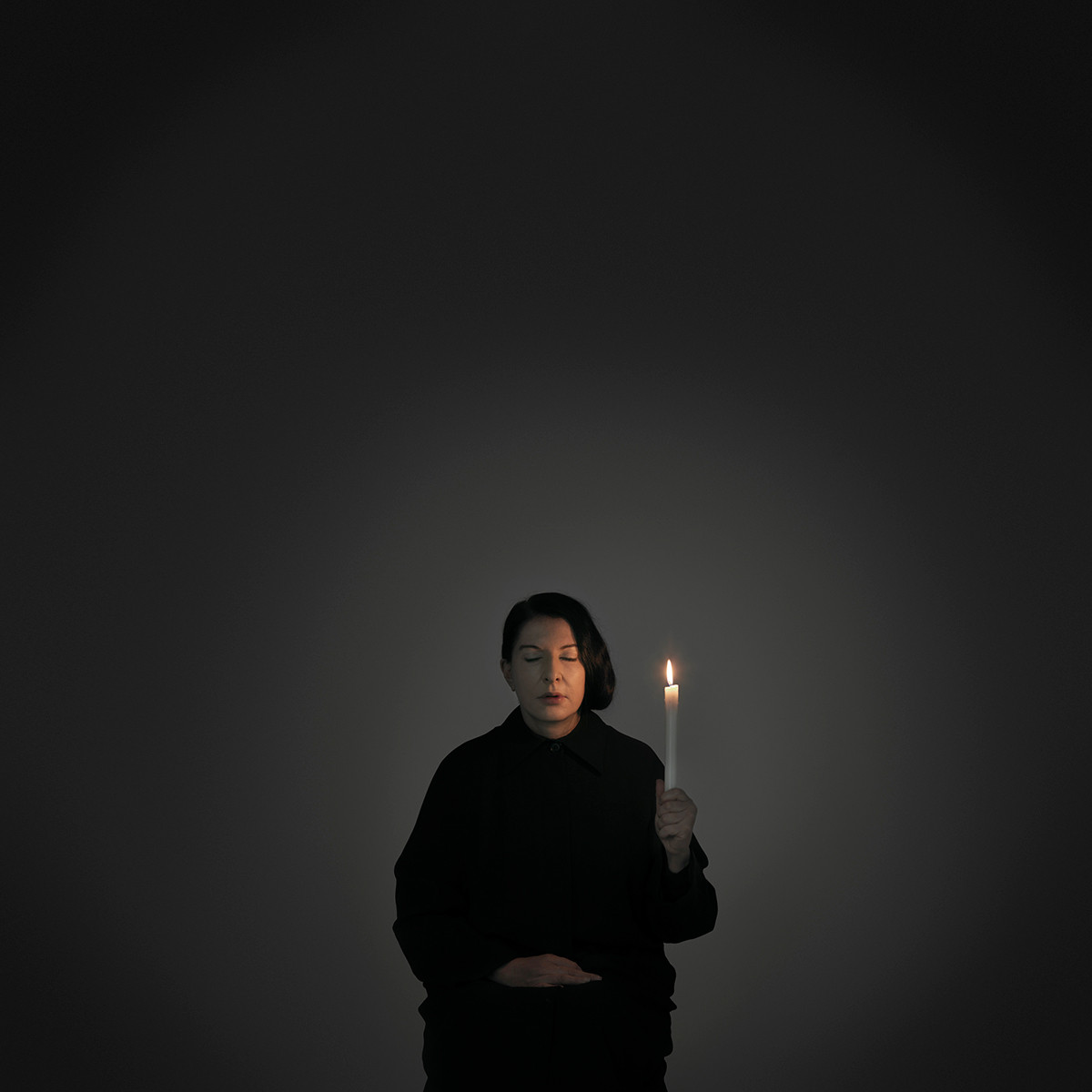 Marina Abramović sits dressed in black and with her eyes closed in a dark room, lit only by the candle in her hand.