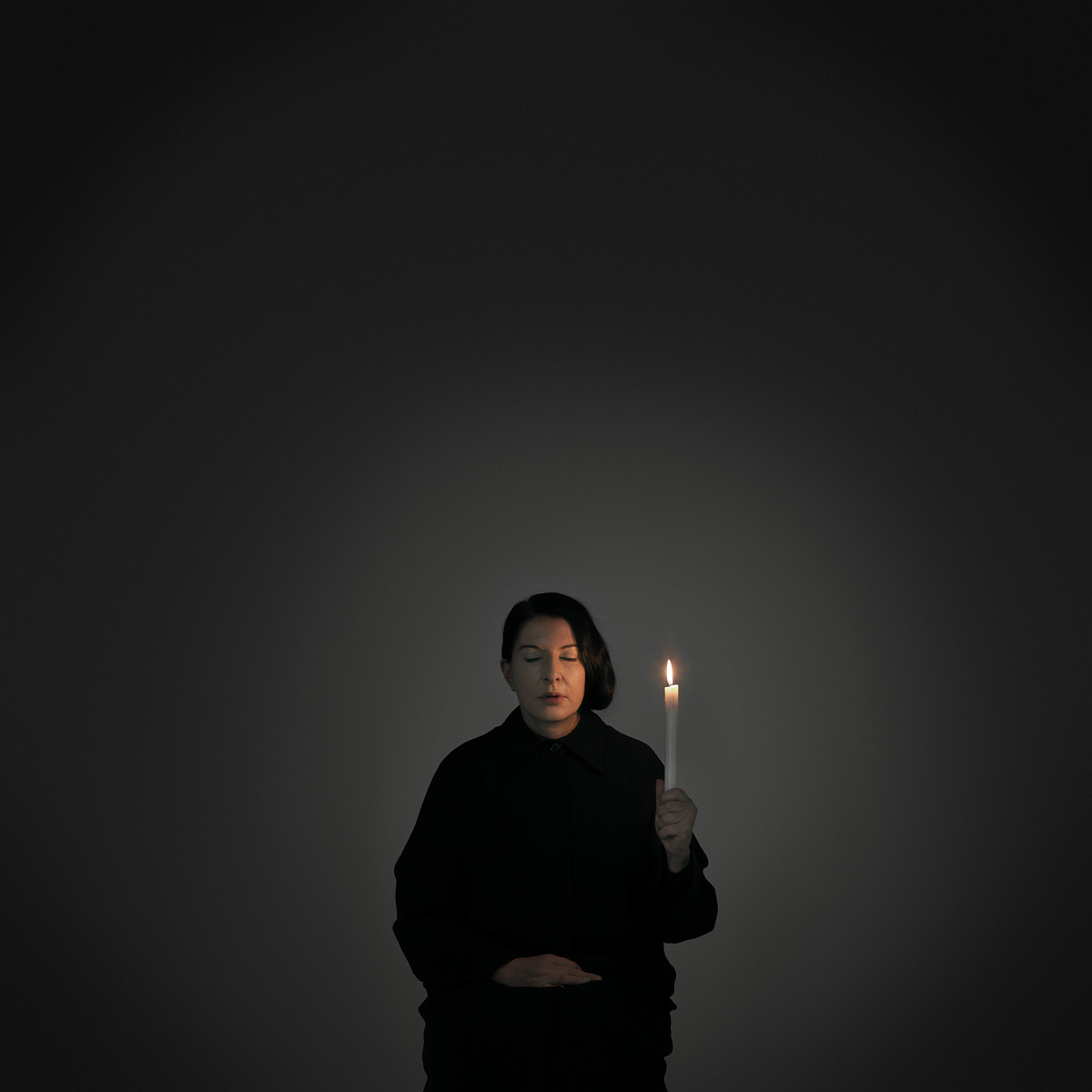 Marina Abramović sits dressed in black and with her eyes closed in a dark room, lit only by the candle in her hand.
