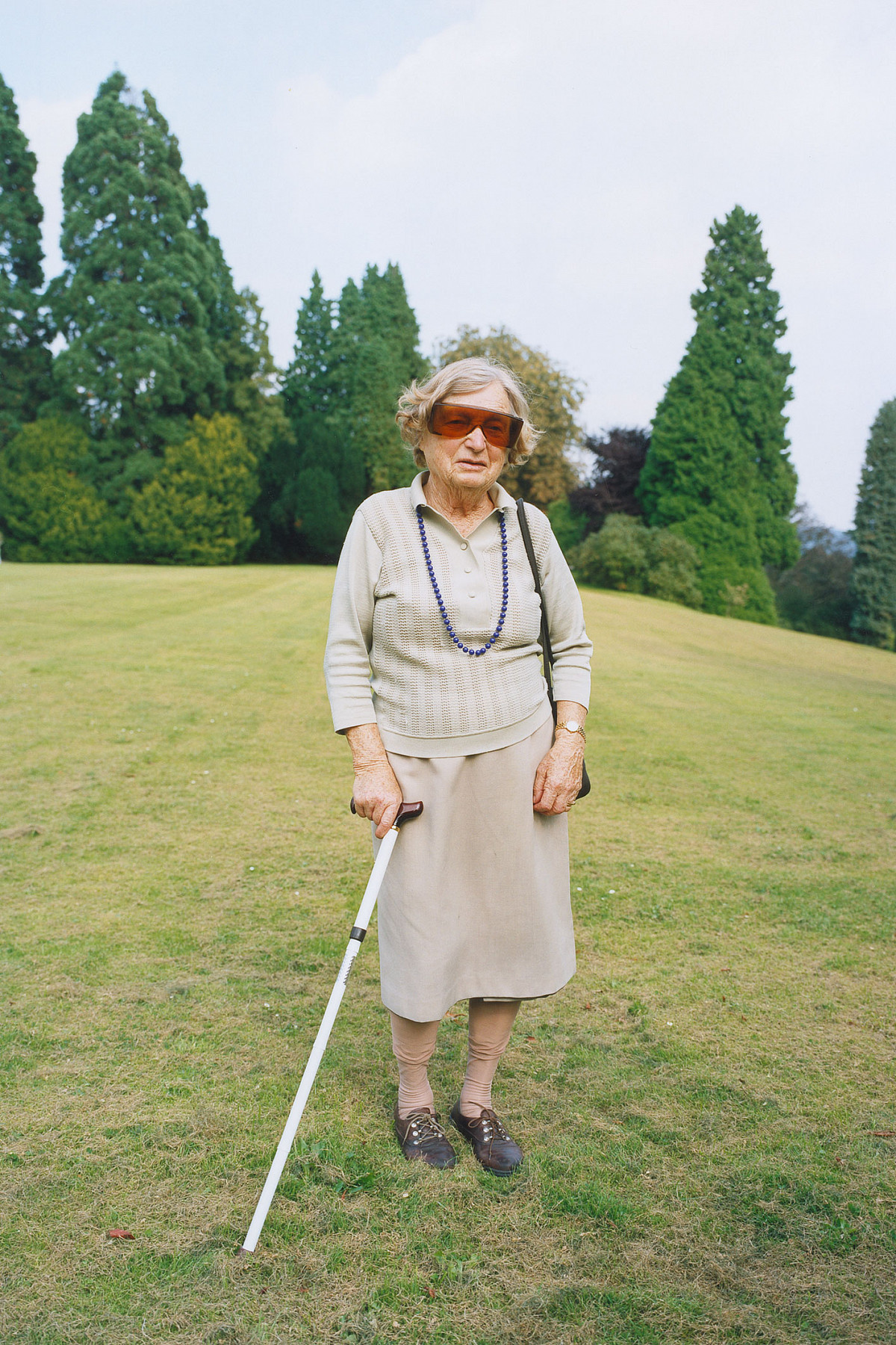 An old woman with a walking stick and large sunglasses stands on a freshly mown meadow.