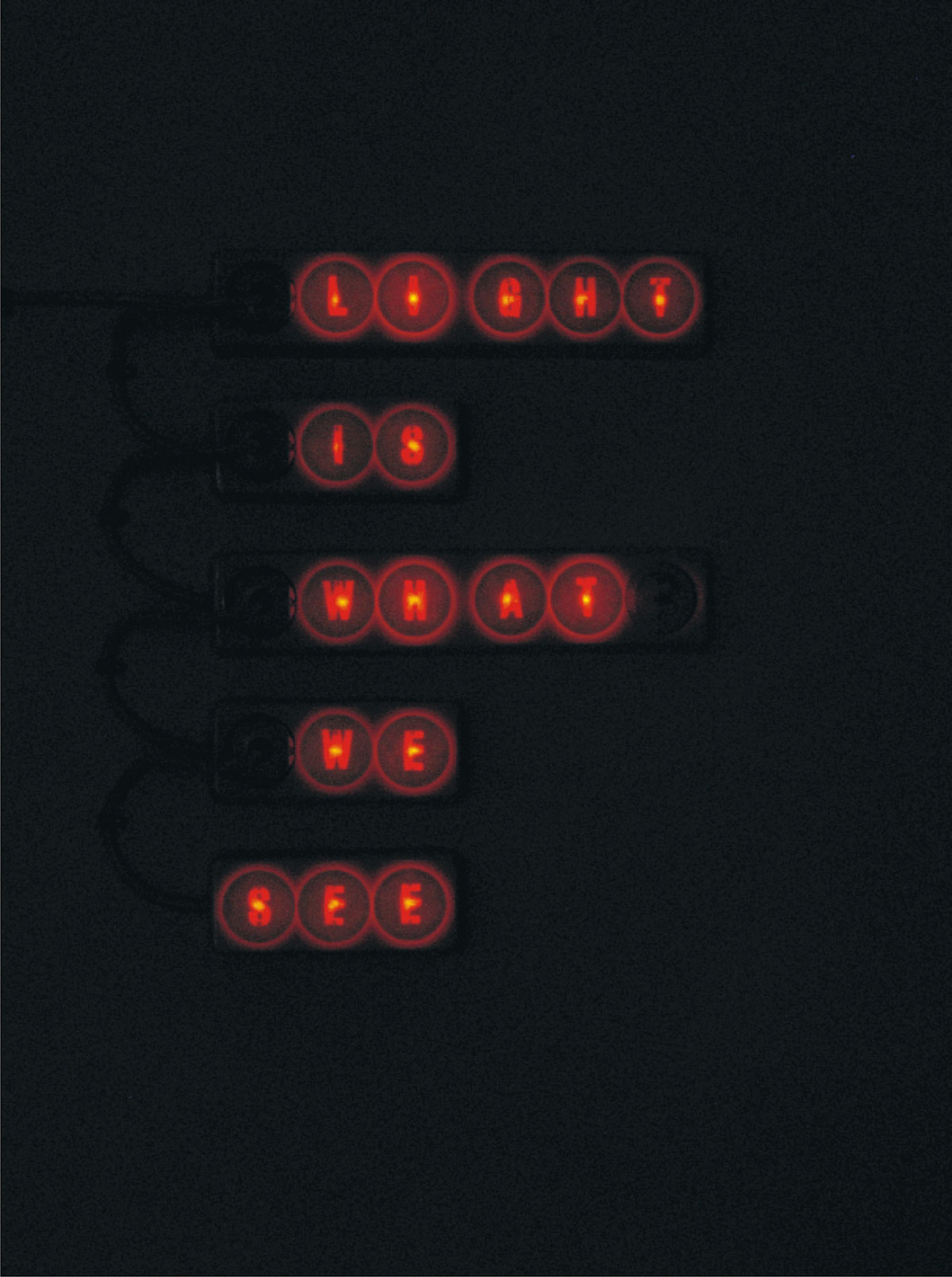 Installation in the dark. The sentence “Light is what we see” appears in red light. The words are arranged one below the other.