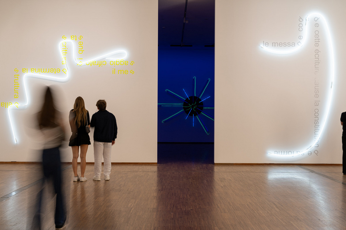 The exhibition hall overlooks a passageway to the next room. The next room is dark and displays a work with neon tubes in the center. On the walls to the left and right are two installations with curved fluorescent tubes surrounded by text written in yellow and white.
