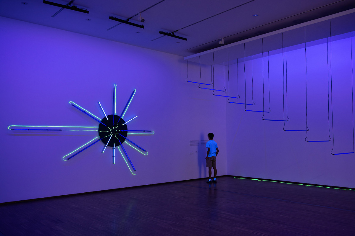 A dark room with neon lighting. On the wall is a circular installation with several neon tubes that spread out from the circle like rays of sunlight. On the other side of the wall, fluorescent tubes hang from the ceiling like a flying staircase.