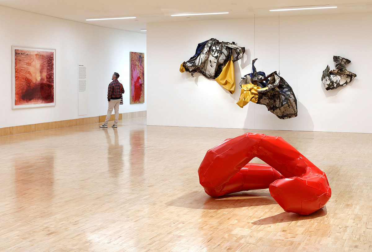 View of the exhibition hall with a red curved sculpture on the floor and abstract works on the walls.