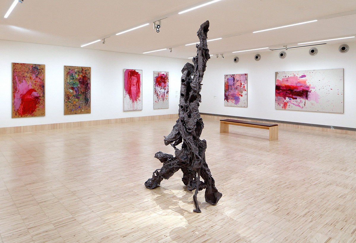 View of the exhibition hall with an abstract, solid sculpture in the center and colorful paintings on the walls.