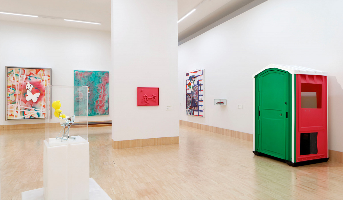 In the exhibition hall: on the left, a display case with a yellow teddy bear on an iron frame; in the middle, a pillar with a red painting; on the right, a mobile toilet cubicle in green, red, and white | Credit: Photo © Rainer Iglar