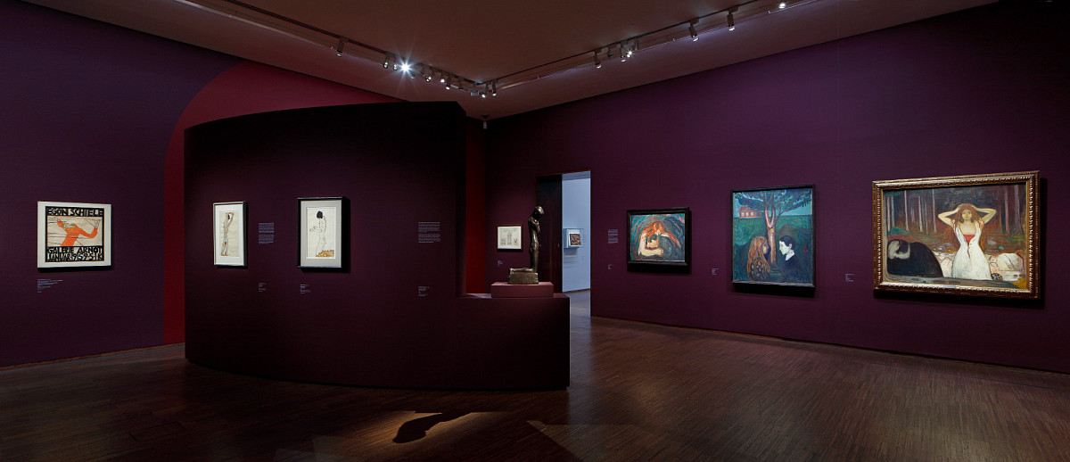 A room in the exhibition with dark red walls and paintings on the walls. In the middle of the room is a wall installation with paintings attached to it and a sculpture standing on a pedestal.