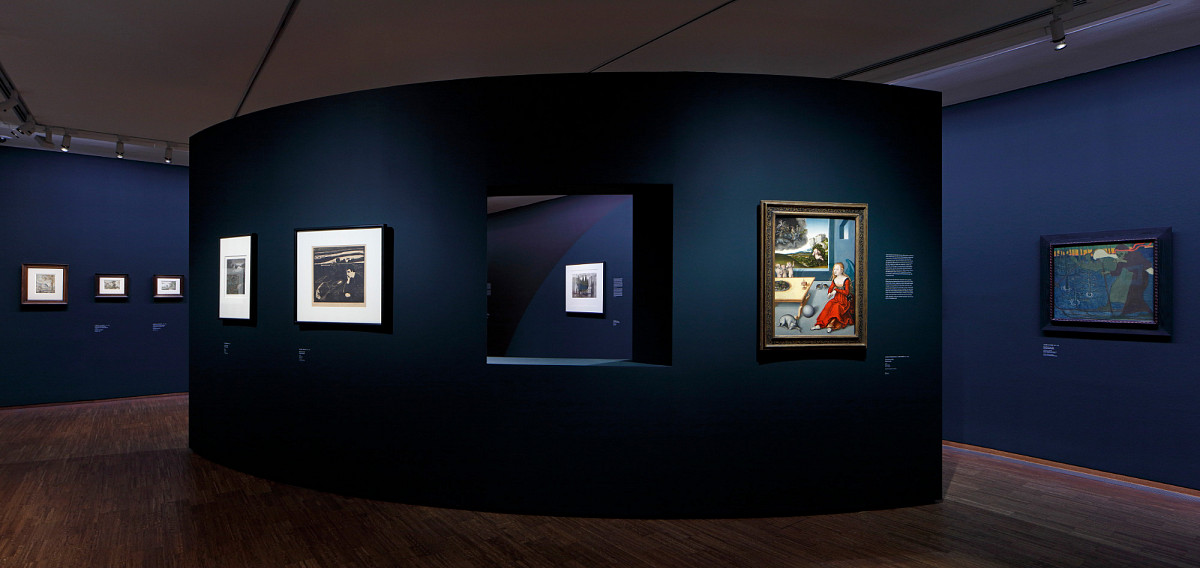 An exhibition room with dark blue walls and a slightly curved wall in the middle with a window through which you can see a painting on the wall behind it.