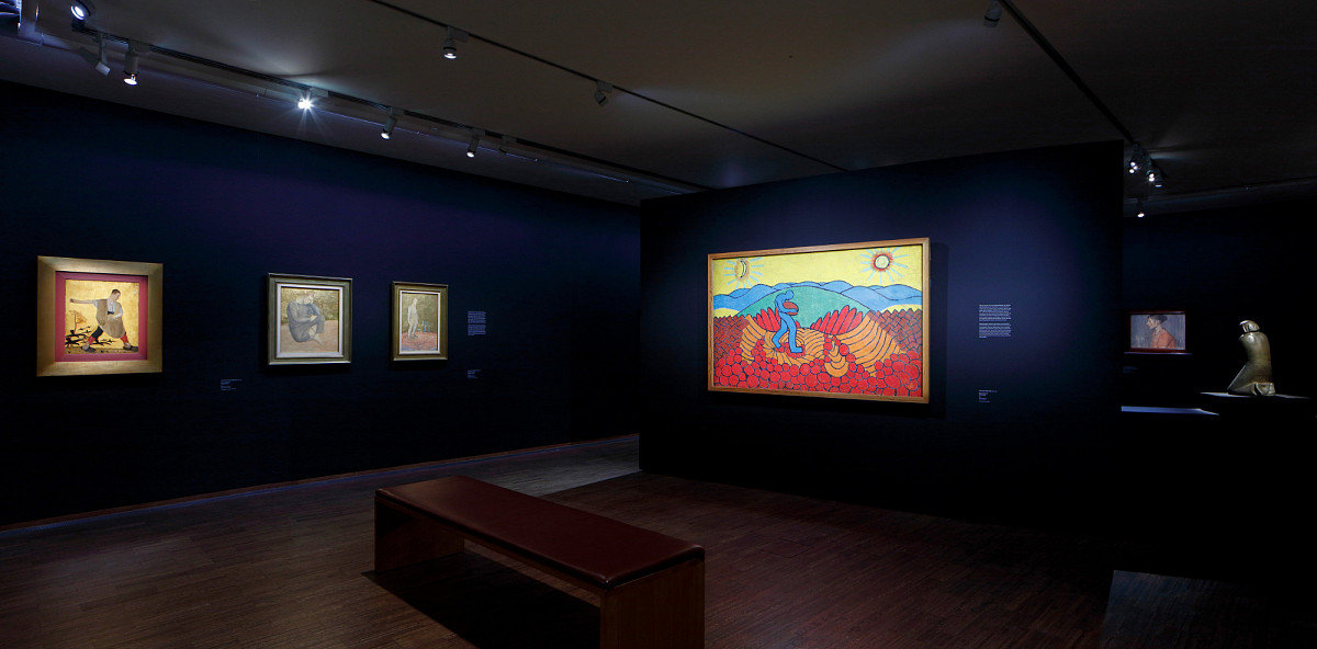 An exhibition room with dark blue walls and a bench in the middle. A large painting in bright colors on one wall. Three smaller paintings, also in bright colors, on the wall next to it.