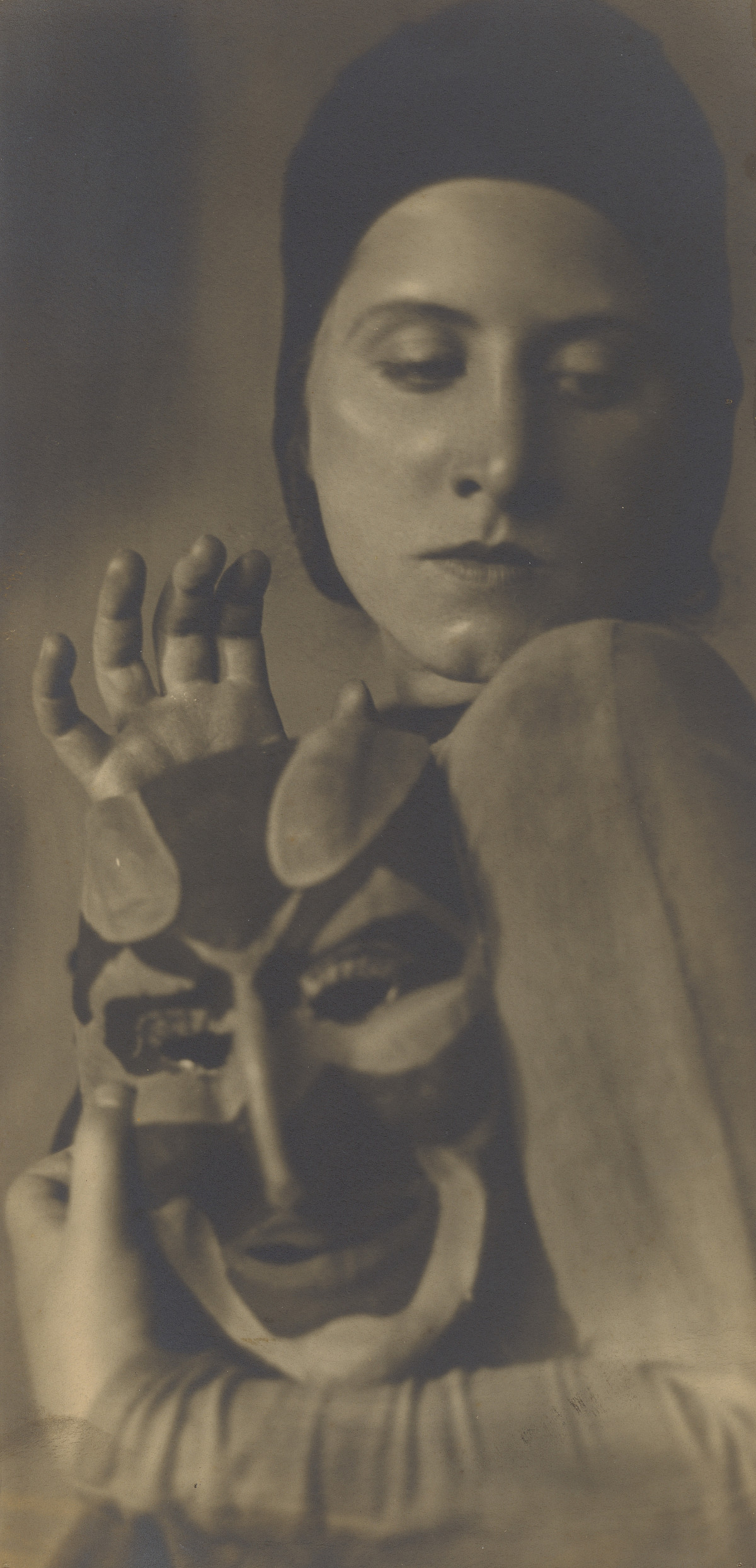 Photograph of dancer Hilde Holger holding a mask in her hand, her head bowed and gaze turned to one side, one hand raised above the mask.
