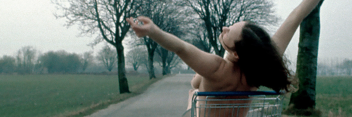 Photograph: A woman sits naked in a shopping cart and stretches her arms into the air. The shopping cart is parked on an empty country road. Fields and an avenue lined with bare trees can be seen to the left and right. Credit: Kirsten Justesen | Lunch, 1975/2018 | © Kirsten Justesen / Courtesy VERBUND COLLECTION, Vienna © Kirsten Justesen / Bildrecht, Vienna 2026