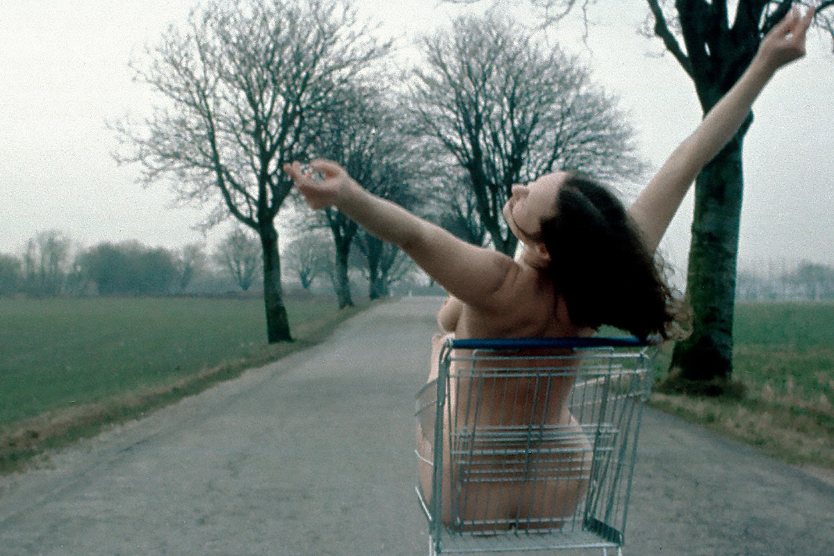 Photograph: A woman sits naked in a shopping cart and stretches her arms into the air. The shopping cart is parked on an empty country road. Fields and an avenue lined with bare trees can be seen to the left and right. Credit: Kirsten Justesen | Lunch, 1975/2018 | © Kirsten Justesen / Courtesy VERBUND COLLECTION, Vienna © Kirsten Justesen / Bildrecht, Vienna 2026
