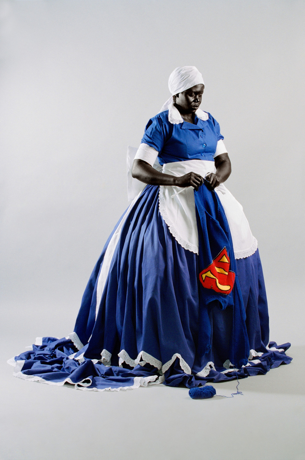A woman in an opulent white and blue maid's costume, knitting a Superman cape.