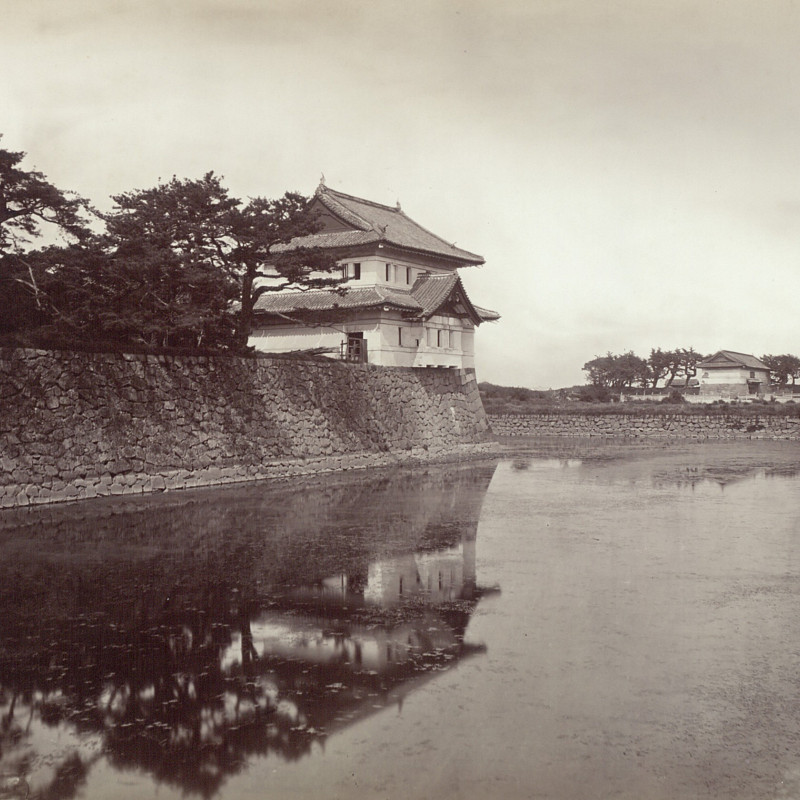 Schwarz-Weiß-Fotografie eines Wachgebäudes im japanischen Stil am Rande des Burggrabens und von Bäumen umgeben.