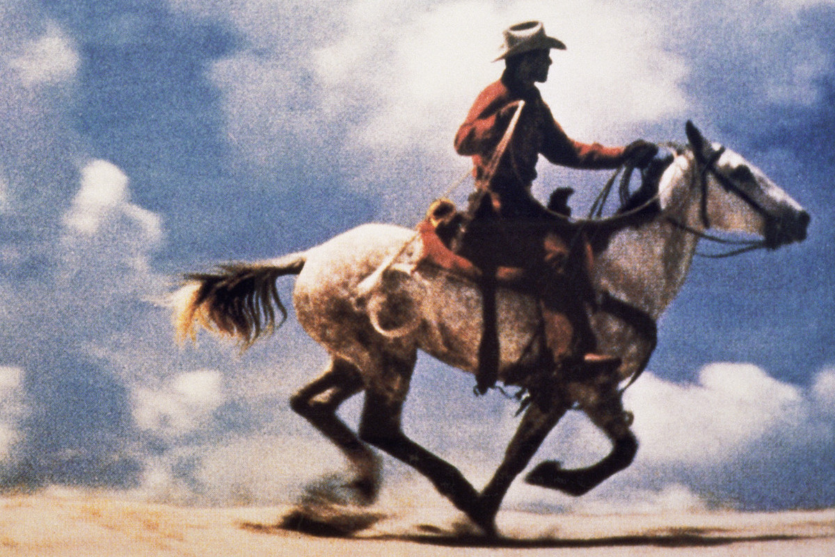 Color photograph of a cowboy riding a galloping horse in an open landscape; large-scale, horizontal composition.