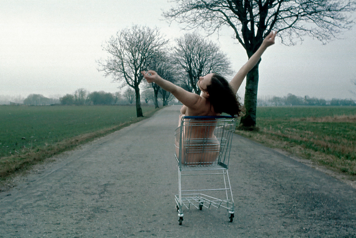 Photograph: A woman sits naked in a shopping cart and stretches her arms into the air. The shopping cart is parked on an empty country road. Fields and an avenue lined with bare trees can be seen to the left and right.