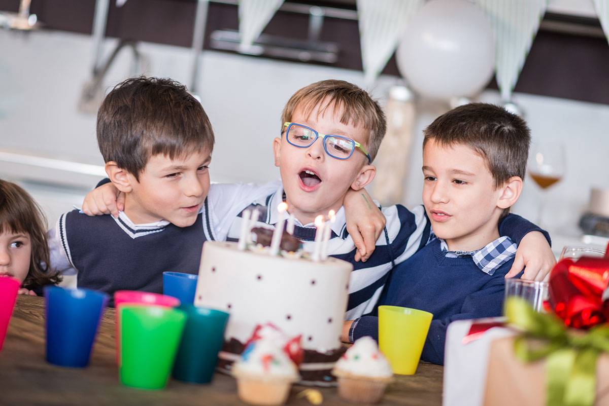 Three children hugging each other. In the middle a boy with glasses, blowing out the candles on the birthday cake. | Photo © ilsbusca